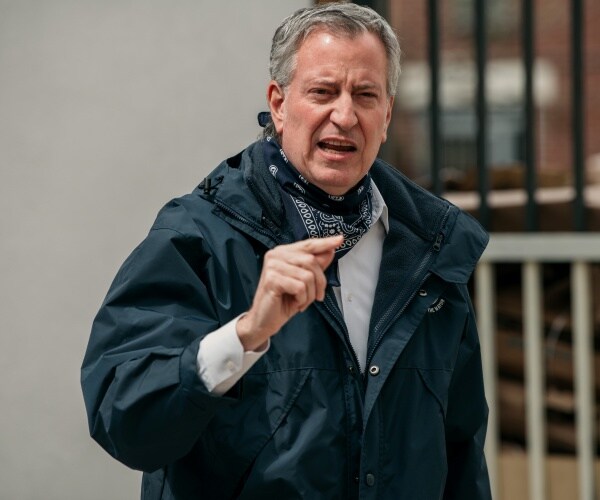 de blasio in a black windbreaker jacket and bandana around his neck standing outside