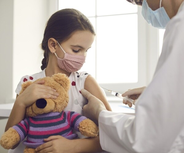 young girl in mask holding teddy bear getting COVID vaccine