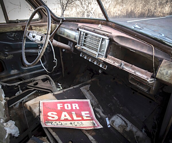 a for sale sign sits on the front seat bench of an old rusted out car at a junk yard