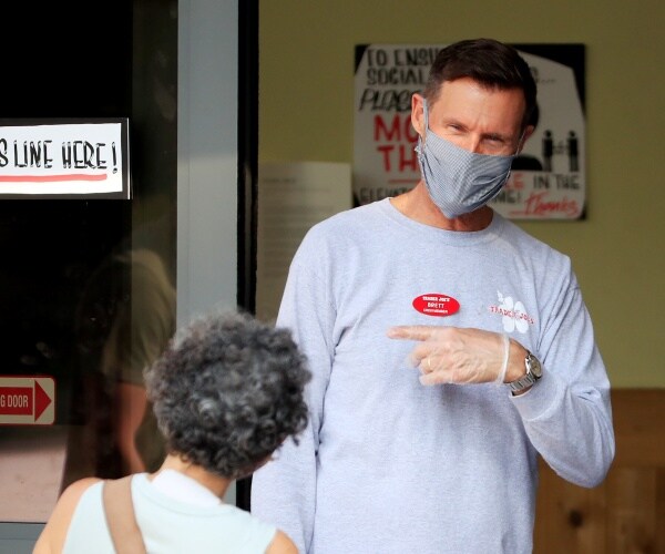 trader joes employee wearing a gray long sleeve shirt and a mask talks to a woman coming in