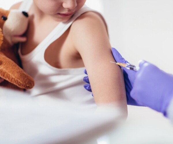 young boy holds teddy bear as a masked healthcare worker gives him a shot