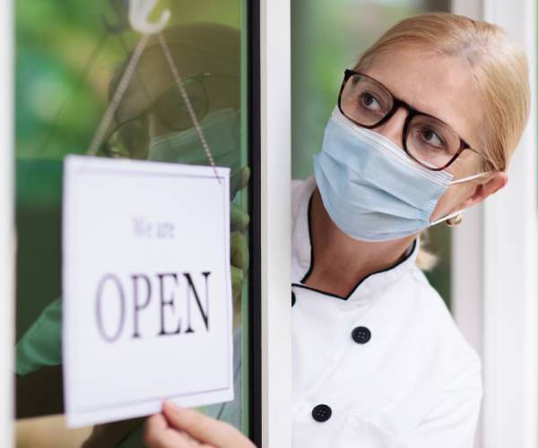 a shopkeeper in a mask with an open sign on her store
