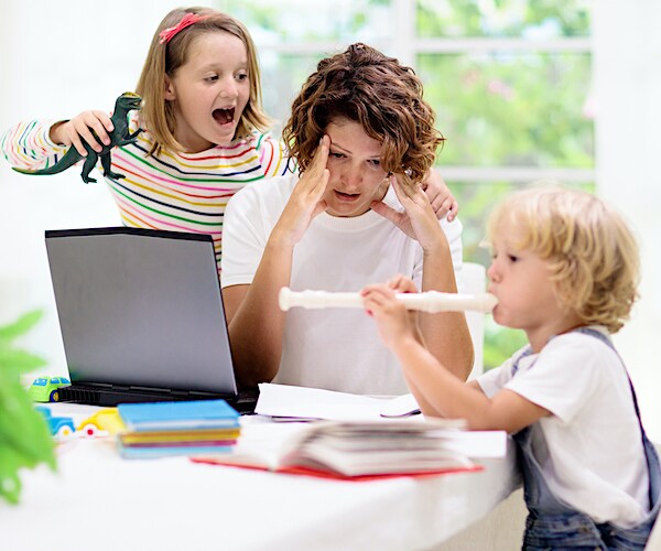 a woman on a laptop massages her temples with kids exulting around her