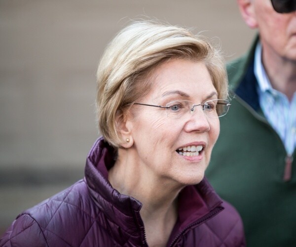 warren in a purple jacket speaking outside with a man in a green jacket behind her