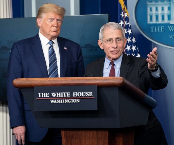 fauci in a black suit speaking with trump standing beside in a navy blue suit and striped tie