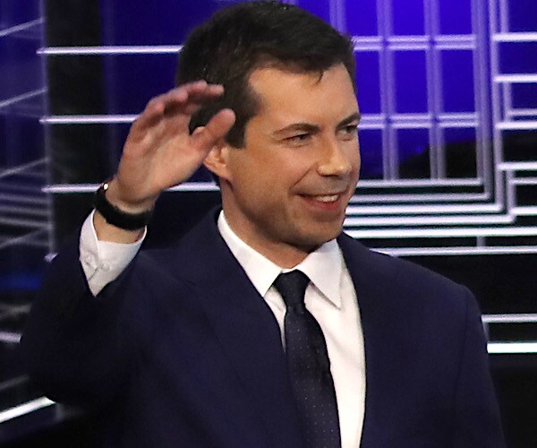pete buttigieg raises his right hand and smiles during a democratic presidential primary debate