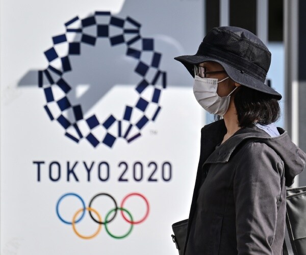 woman in a mask and a black hat walks by a tokyo 2020 poster