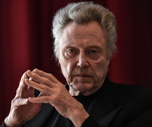 christopher walken sits with hands steepled in front of red curtain
