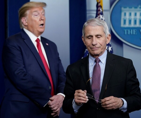 trump in a suit and red tie standing behind fauci in a light blue shirt and purple tie with his glasses off