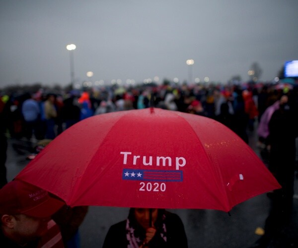 woman under a red trump 2020 umbrella with a crowd of trump supporters waiting outside in the background