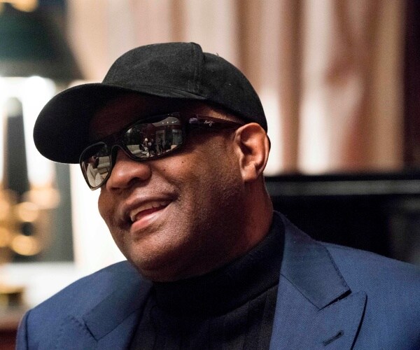 bell wears a blue suit and black shirt, sunglasses, and a baseball cap