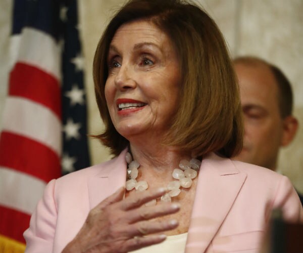 nancy pelosi wearing a pink suit with white shell blouse smiles as she gestures with her right hand