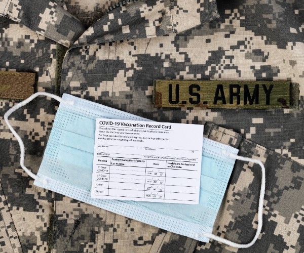 an army uniform with a covid vaccine card and a surgical mask lying on it