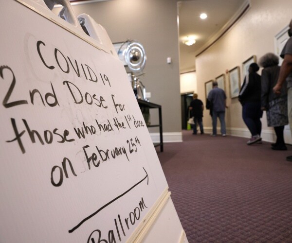 people line up on a hall with a vaccine sign in the foreground.