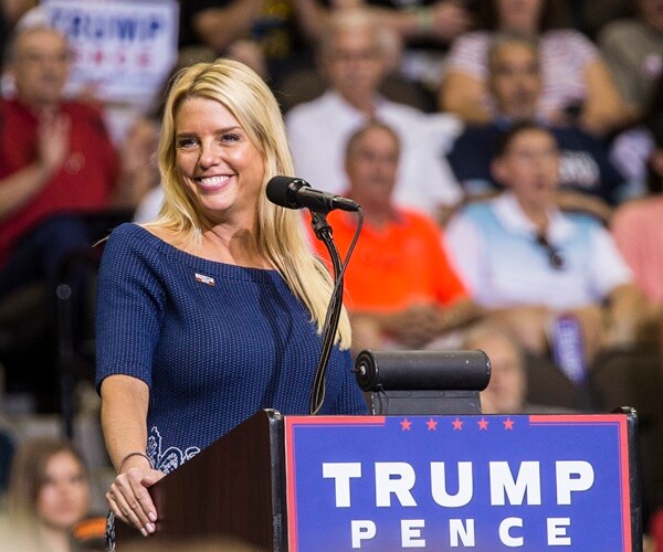 pam bondi stands at a podium at a rally with a trump pence sign on it