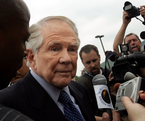 robertson standing in a crowd wearing a suit and blue printed tie with a mic held up to him