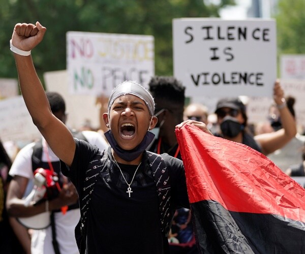 young man in black raises a fist with protesters holding signs in the background