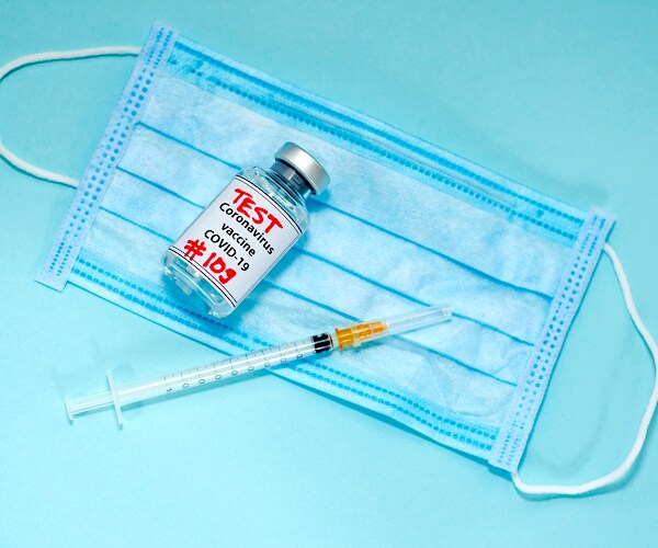 a facemask lays on a table with a vile marked covid-19 test and a syringe with a needle
