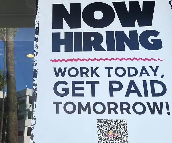 now hiring sign hangs in window