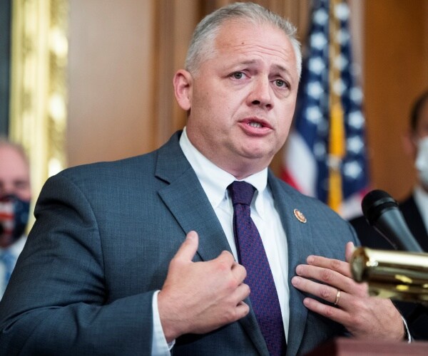 denver riggleman speaks at a mic in front of a flag and masked men