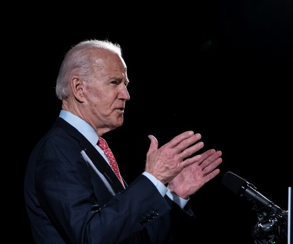 biden in a suit and blue shirt and red tie speaking into a mic