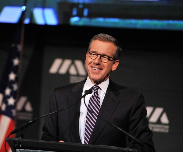 brian williams in a black suit and striped tie speaking at a podium with an american flag in the background