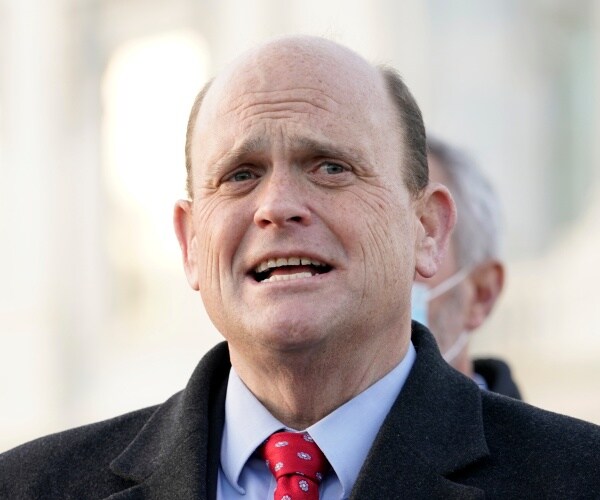 reed in a black coat and red tie speaking outside