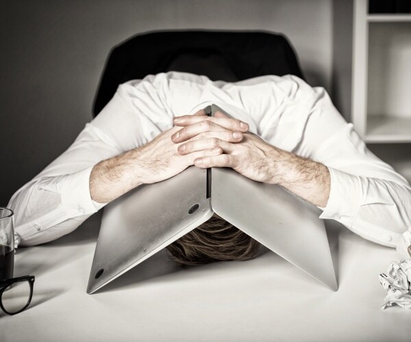 man at work suffering from burnout with laptop on head, papers crumpled on side