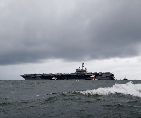 uss ronald reagan is shown with cloudy skies and a wave rolling by in front