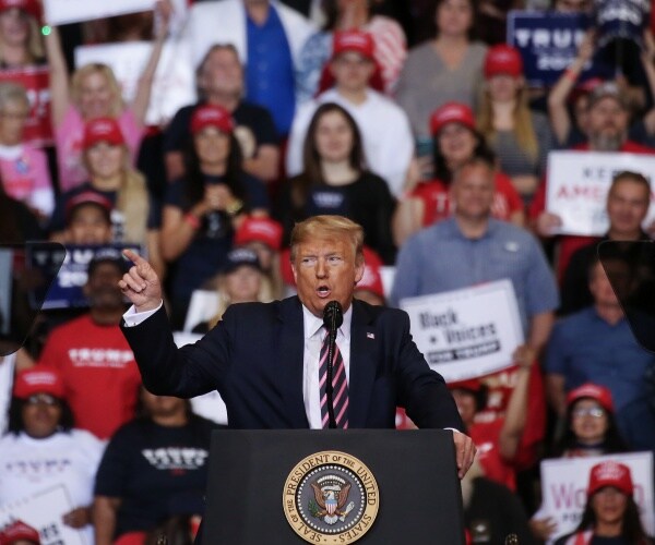 trump in a navy blue suit and pink and black striped tie speaking at a rally