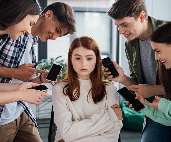 teenaged girl sits on chair as peers are all around her showing her their smartphones