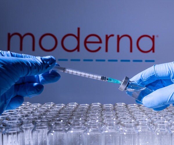 gloved hand holding a vial of Moderna COVID vaccine and a syringe, and Moderna logo in background
