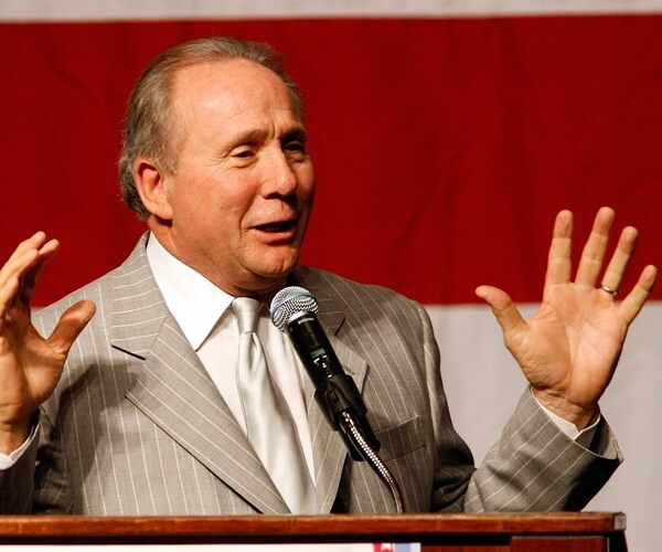 michael reagan speaks standing in front of a flag