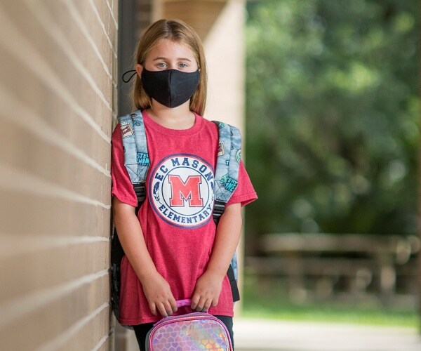 little girl leans on building wearing mask and holding lunch box