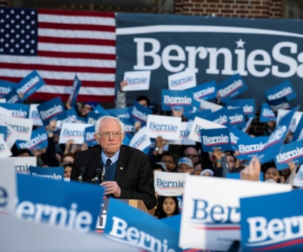 bernie sanders speaks during a rally to many supporters holding up bernie signs with an american flag behind him