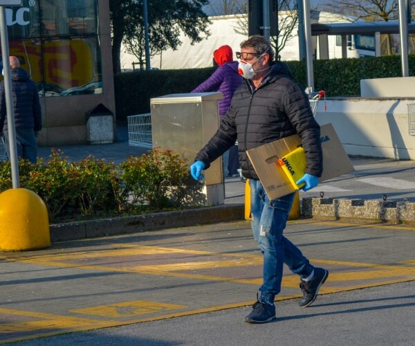 man in a balck jacket and blue gloves wears a mask while walking in a parking lot