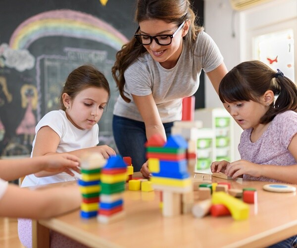 preschool students sits at table with blocks and teacher stands over them