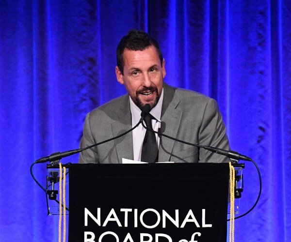adam sandler in a gray suit and black tie speaks in front of blue velvet curtains