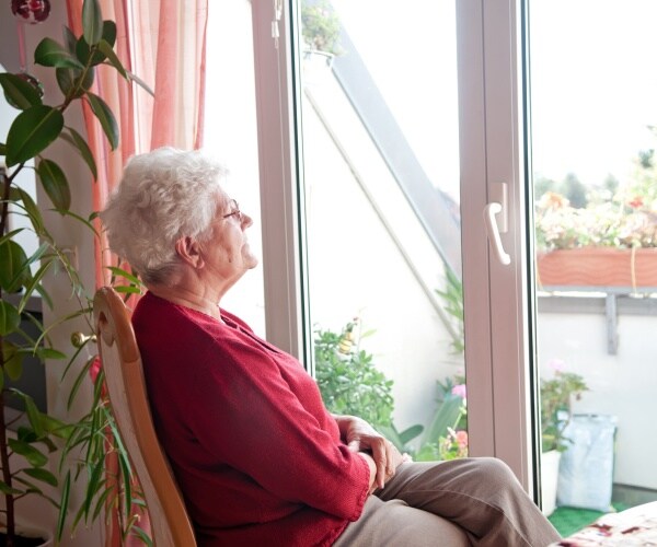 old woman sitting in a chair and looking outside a glass door wearing a red top
