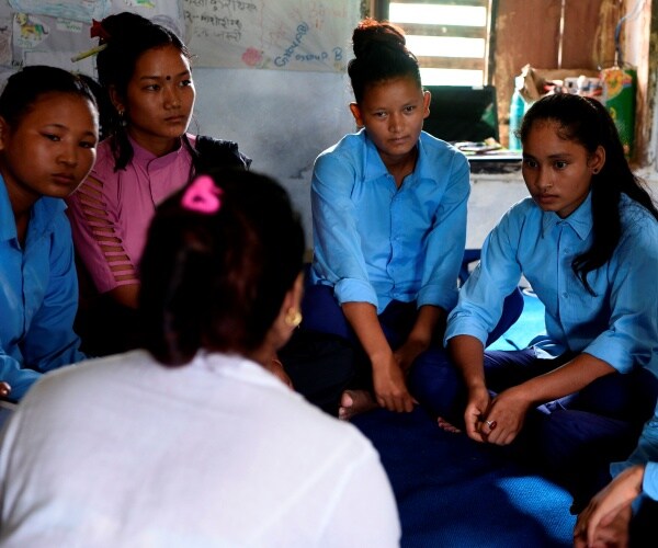 young woman in pink talks to girls dressed in blue sitting in a circle
