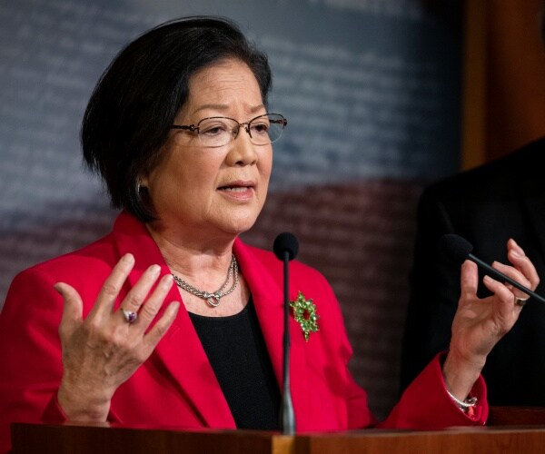 mazie hirono in a bright pinkish red suit and black top speaking at a podium
