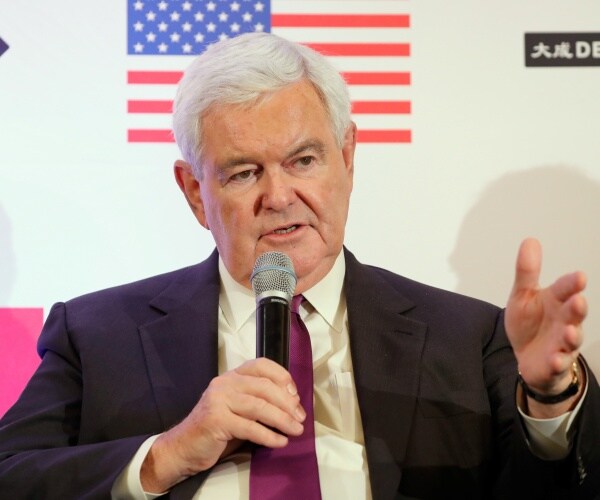 newt gingrich in a suit and purple tie speaking into a mic with an american flag icon behind him