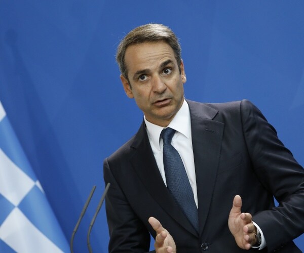 mitsotakis in a black suit and blue tie speaking next to a greek flag