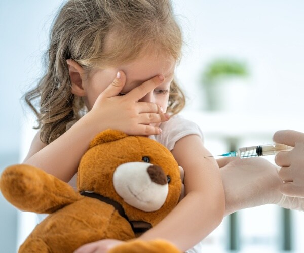 young girl covering her eyes, holding teddy bear, while getting a vaccine