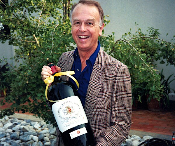 Trade Joe's founder Joe Coulombe smiles holding a large bottle of wine
