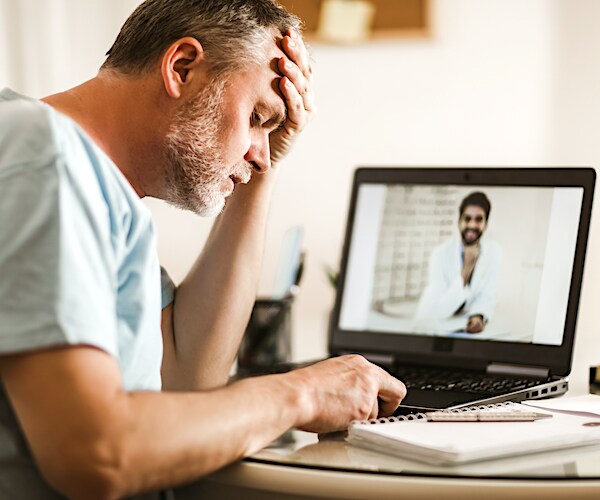 a man buries his head in his left hand while on a video conference with his smiling doctor