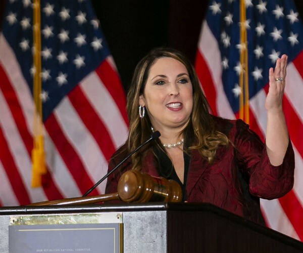 ronna mcdaniel is shown in a burgundy suit with the us flag in the background