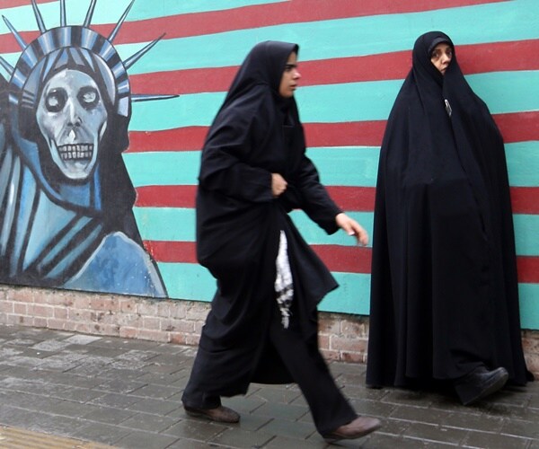 women in hijabs walk past anti-american murals in iran