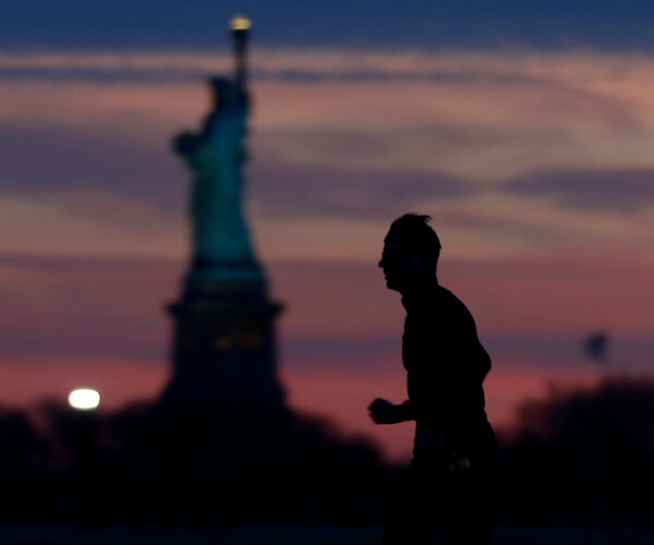 a male jogger in the foreground shadows runs with the statue of liberty towering in the background