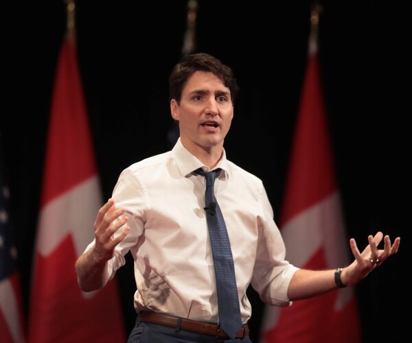 justin trudeau in a white shirt and blue tie speaking in front of american and canadian flags